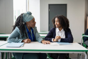 How to choose the right tutor for your child. A supportive female tutor in her early 50s sitting at a desk with a teenage schoolgirl while discussing an assignment.