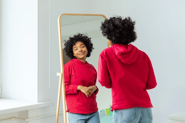 A Black teen girl with curly Afro hair looking admiringly at herself in a mirror with confidence.